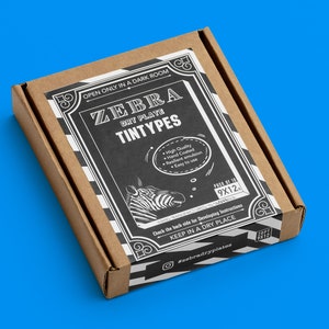 Op de afbeelding: Een bruine kartonnen doos met een zwart-wit label waarop "ZEBRA DRY PLATE TINTYPES" staat. Het label heeft een zebra-illustratie en de tekst "OPEN ONLY IN A DARK ROOM", "PACK OF 10 9X12", "Check the back side for Developing Instructions", "KEEP IN A DRY PLACE" en "#zebradryplates".