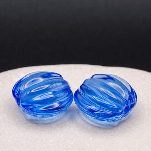 May include: Two translucent blue glass beads with a rounded, ribbed, and pumpkin-like shape. The beads feature a glossy finish and a series of vertical grooves that create a textured surface, reflecting light across their curved, circular forms.