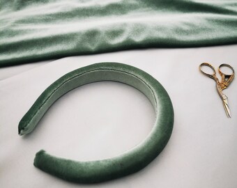 May include: A sage green velvet headband with a simple design. The headband is lying on a white surface.