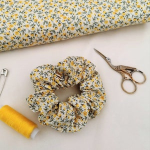 K&ouml;nnte beinhalten: Ein wei&szlig;er Haargummi mit gelbem Blumenmuster. Der Haargummi ist aus Stoff und zu einem Knoten gebunden. Der Haargummi liegt auf einer wei&szlig;en Oberfl&auml;che.