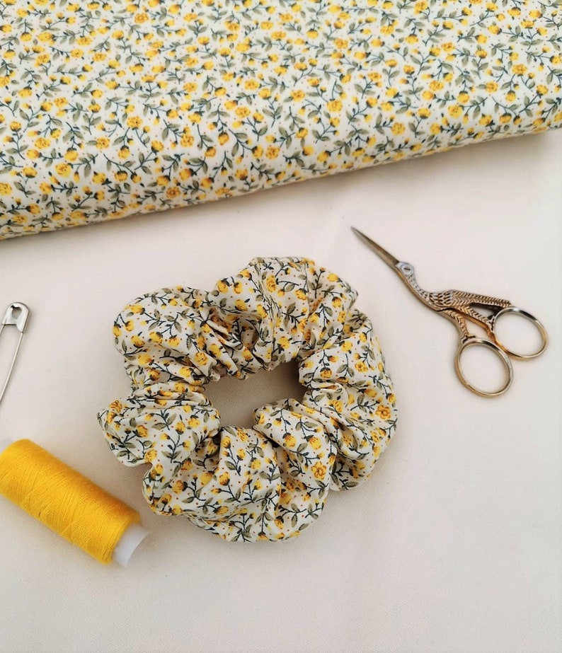K&ouml;nnte beinhalten: Ein wei&szlig;er Haargummi mit gelbem Blumenmuster. Der Haargummi ist aus Stoff und zu einem Knoten gebunden. Der Haargummi liegt auf einer wei&szlig;en Oberfl&auml;che.