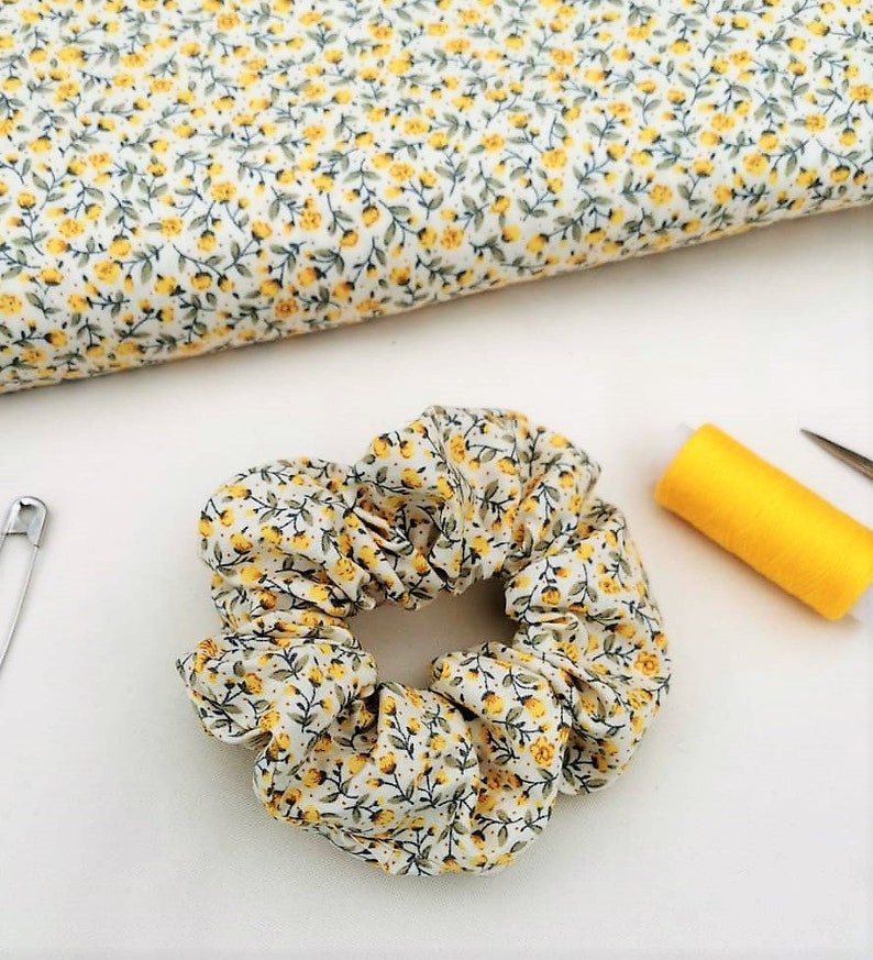 K&ouml;nnte beinhalten: Ein wei&szlig;er Haargummi mit gelbem Blumenmuster. Der Haargummi ist aus Stoff und hat eine gefaltete Optik. Er liegt auf einer wei&szlig;en Oberfl&auml;che.