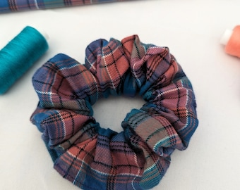 May include: A plaid scrunchie in shades of blue, coral, and purple. The fabric has a classic tartan pattern. A spool of teal thread and a piece of matching fabric are visible in the background.