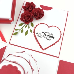 Puede incluir: Una manualidad de papel roja y blanca hecha a mano con tres rosas de papel rojas y hojas verdes. El dise&ntilde;o incluye un elemento en forma de coraz&oacute;n con el texto "f&uuml;r das Traum Paar". Un art&iacute;culo decorativo &uacute;nico.