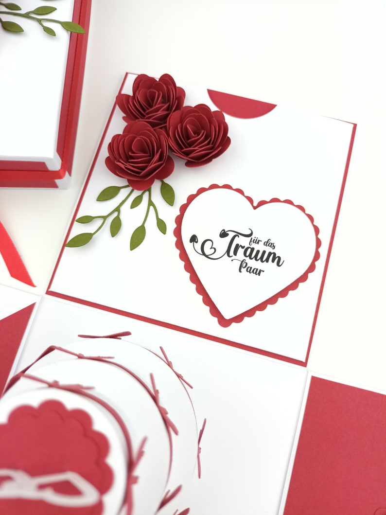Puede incluir: Una manualidad de papel roja y blanca hecha a mano con tres rosas de papel rojas y hojas verdes. El dise&ntilde;o incluye un elemento en forma de coraz&oacute;n con el texto "f&uuml;r das Traum Paar". Un art&iacute;culo decorativo &uacute;nico.