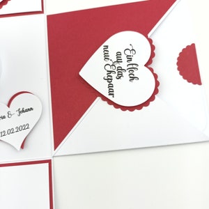 Puede incluir: Tarjeta de felicitaci&oacute;n hecha a mano, blanca y roja, adornada con corazones. La tarjeta incluye una etiqueta en forma de coraz&oacute;n con el texto "Ein Hoch auf das neue Ehepaar" en escritura negra. La tarjeta es para una boda.