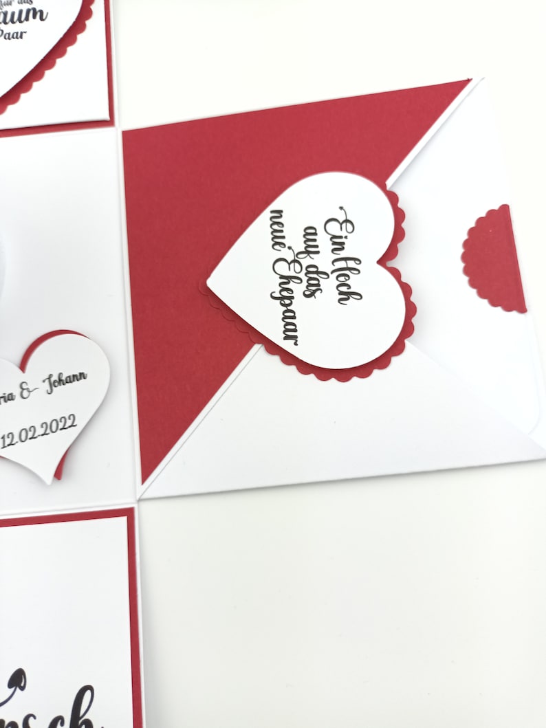 Puede incluir: Tarjeta de felicitaci&oacute;n hecha a mano, blanca y roja, adornada con corazones. La tarjeta incluye una etiqueta en forma de coraz&oacute;n con el texto "Ein Hoch auf das neue Ehepaar" en escritura negra. La tarjeta es para una boda.