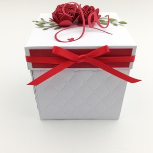 Puede incluir: Una caja de regalo blanca con una cinta roja atada. La caja tiene un patr&oacute;n acolchado en los lados y est&aacute; rematada con rosas de papel rojas, hojas verdes y la palabra "Joy" en escritura roja. Tambi&eacute;n hay un peque&ntilde;o coraz&oacute;n rojo.