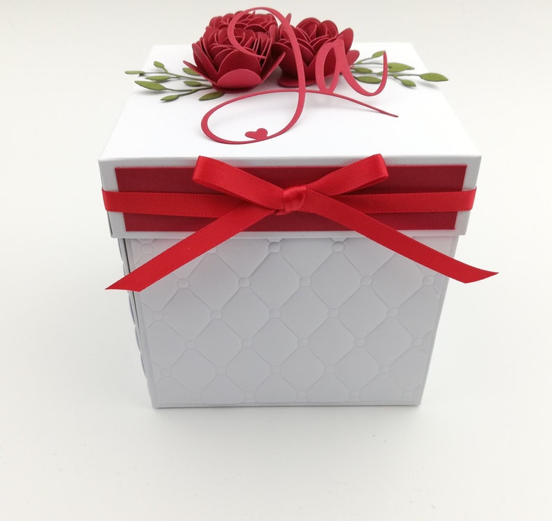 Puede incluir: Una caja de regalo blanca con una cinta roja atada. La caja tiene un patr&oacute;n acolchado en los lados y est&aacute; rematada con rosas de papel rojas, hojas verdes y la palabra "Joy" en escritura roja. Tambi&eacute;n hay un peque&ntilde;o coraz&oacute;n rojo.