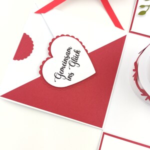 Puede incluir: Una tarjeta de felicitaci&oacute;n blanca y roja con un adorno en forma de coraz&oacute;n. El coraz&oacute;n tiene el texto "Gemeinsam ins Gl&uuml;ck" en escritura negra. Una cinta roja est&aacute; atada alrededor de una caja blanca en el fondo.