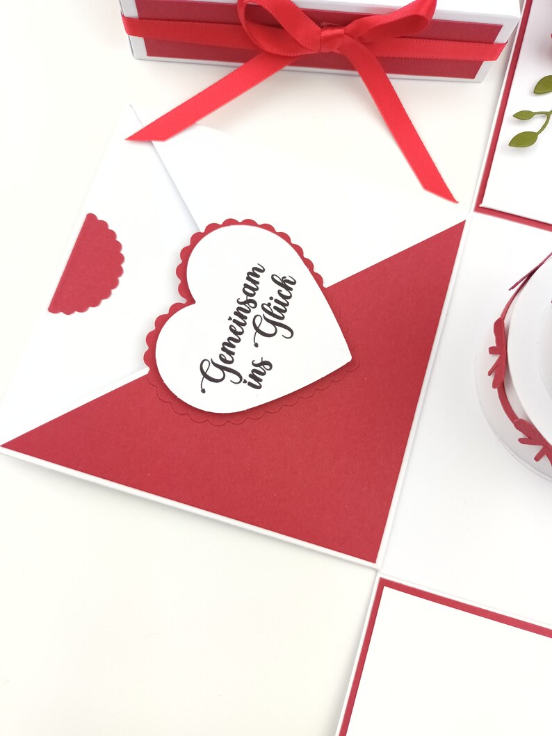 Puede incluir: Una tarjeta de felicitaci&oacute;n blanca y roja con un adorno en forma de coraz&oacute;n. El coraz&oacute;n tiene el texto "Gemeinsam ins Gl&uuml;ck" en escritura negra. Una cinta roja est&aacute; atada alrededor de una caja blanca en el fondo.