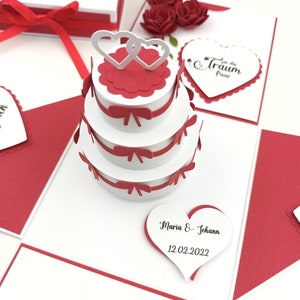 Puede incluir: Una caja explosiva de tem&aacute;tica nupcial blanca y roja con una pieza central de pastel de tres niveles. El pastel tiene detalles en rojo y dos adornos de coraz&oacute;n entrelazados. La caja incluye tarjetas en forma de coraz&oacute;n y rosas rojas decorativas.