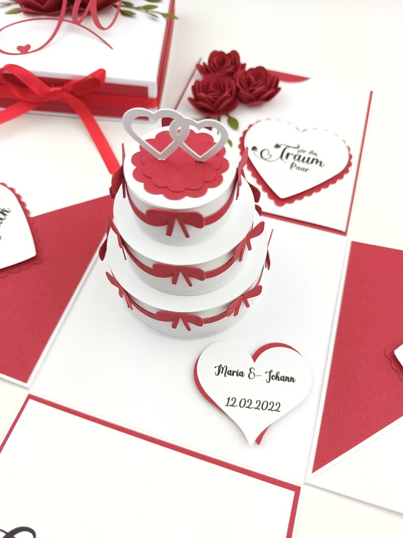 Puede incluir: Una caja explosiva de tem&aacute;tica nupcial blanca y roja con una pieza central de pastel de tres niveles. El pastel tiene detalles en rojo y dos adornos de coraz&oacute;n entrelazados. La caja incluye tarjetas en forma de coraz&oacute;n y rosas rojas decorativas.