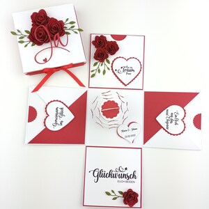 Puede incluir: Una caja explosiva blanca y roja con rosas de papel y hojas decorativas. La caja abierta revela m&uacute;ltiples compartimentos con detalles en forma de coraz&oacute;n y texto en alem&aacute;n. Una cinta roja cierra la caja.