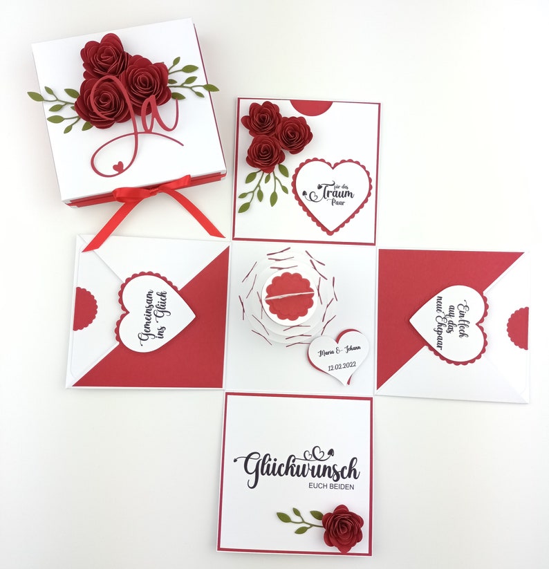 Puede incluir: Una caja explosiva blanca y roja con rosas de papel y hojas decorativas. La caja abierta revela m&uacute;ltiples compartimentos con detalles en forma de coraz&oacute;n y texto en alem&aacute;n. Una cinta roja cierra la caja.