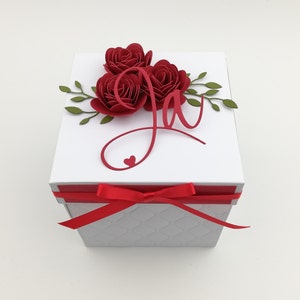 Puede incluir: Una caja de regalo blanca con una cinta y un lazo rojos. La parte superior est&aacute; adornada con tres rosas de papel rojas, hojas verdes y una letra "J" cursiva roja con un coraz&oacute;n. La caja tiene un patr&oacute;n acolchado.