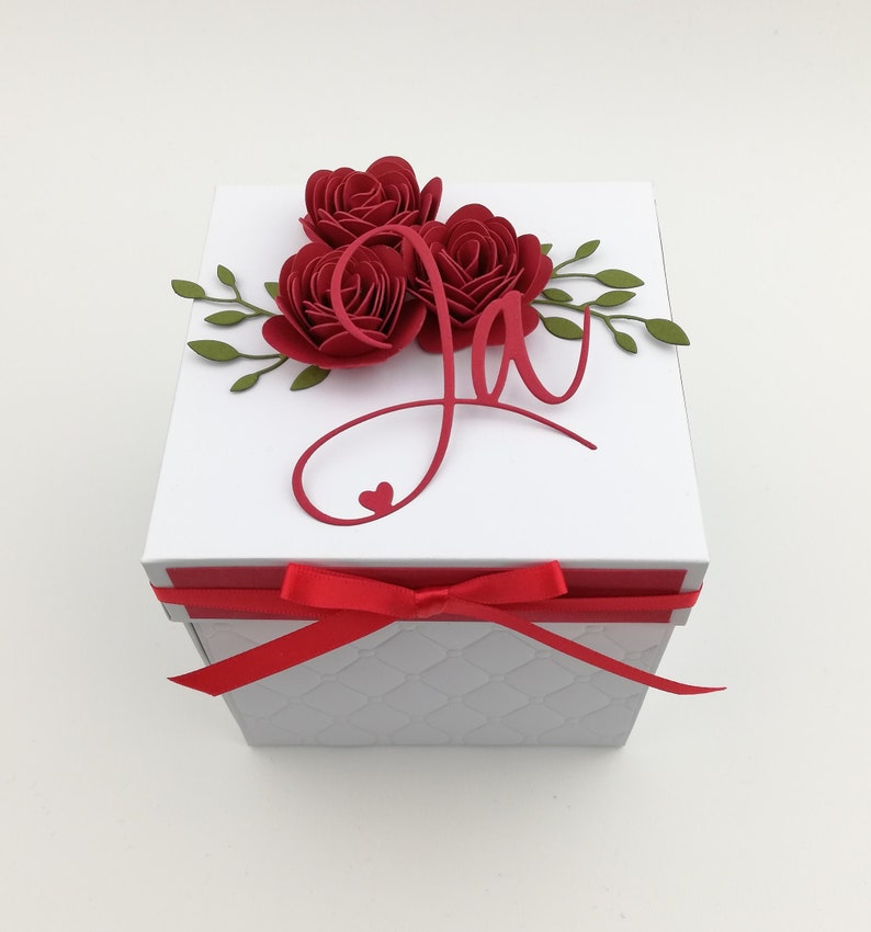 Puede incluir: Una caja de regalo blanca con una cinta y un lazo rojos. La parte superior est&aacute; adornada con tres rosas de papel rojas, hojas verdes y una letra "J" cursiva roja con un coraz&oacute;n. La caja tiene un patr&oacute;n acolchado.