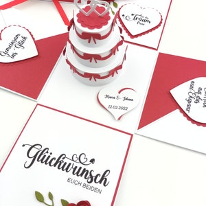 Puede incluir: Tarjeta de boda hecha a mano, blanca y roja. Presenta una tarta de tres pisos, decoraciones en forma de coraz&oacute;n y las palabras "Gl&uuml;ckwunsch euch beiden". Tambi&eacute;n se ven rosas rojas decorativas y una caja de regalo con una cinta roja. Incluye la fecha 12.02.2022.