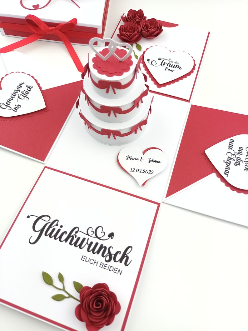 Puede incluir: Tarjeta de boda hecha a mano, blanca y roja. Presenta una tarta de tres pisos, decoraciones en forma de coraz&oacute;n y las palabras "Gl&uuml;ckwunsch euch beiden". Tambi&eacute;n se ven rosas rojas decorativas y una caja de regalo con una cinta roja. Incluye la fecha 12.02.2022.