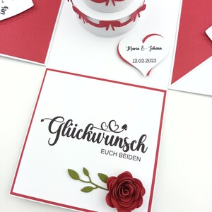 Puede incluir: Tarjeta de boda hecha a mano con una tarta desplegable de tres pisos y lazos rojos. La tarjeta presenta las palabras "Gl&uuml;ckwunsch Euch Beiden" y una rosa de papel roja. Una etiqueta en forma de coraz&oacute;n dice "Maria & Johann 12.02.2022".