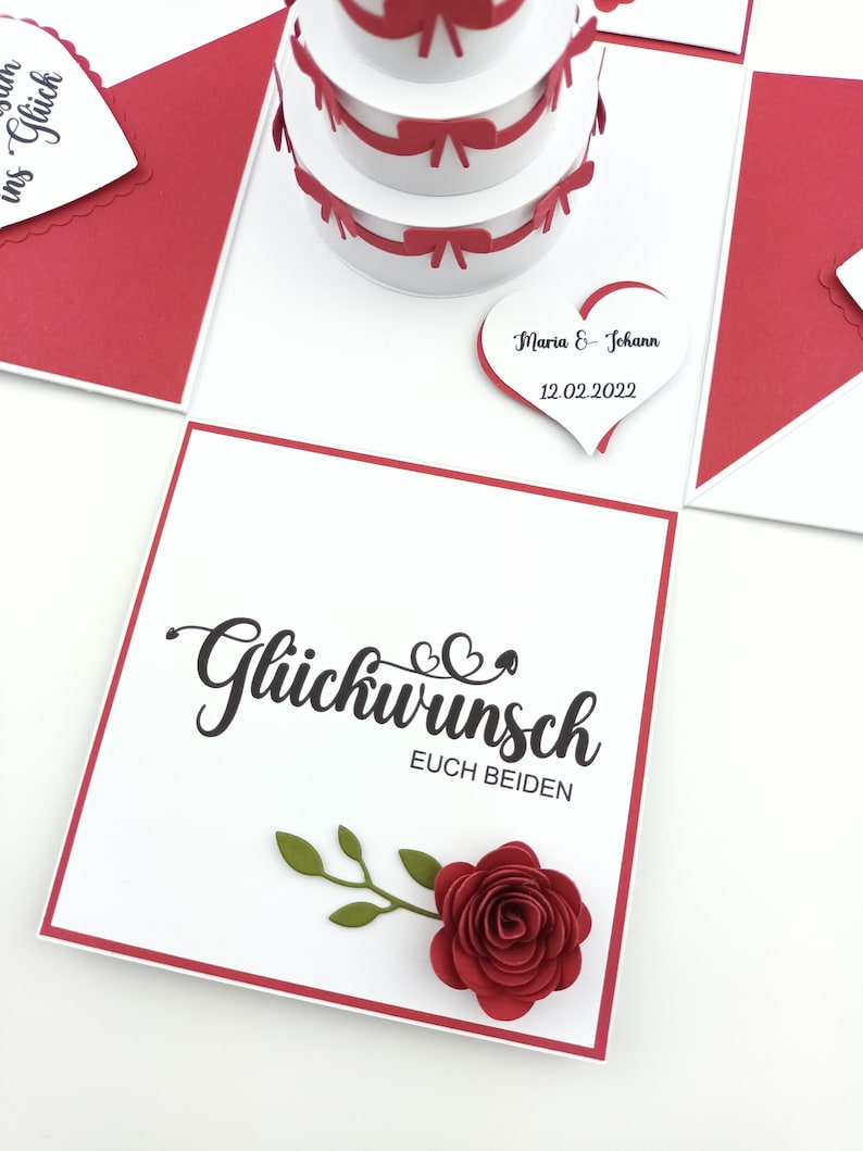 Puede incluir: Tarjeta de boda hecha a mano con una tarta desplegable de tres pisos y lazos rojos. La tarjeta presenta las palabras "Gl&uuml;ckwunsch Euch Beiden" y una rosa de papel roja. Una etiqueta en forma de coraz&oacute;n dice "Maria & Johann 12.02.2022".