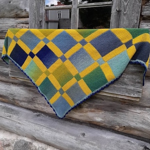 Könnte beinhalten: Eine gelbe, blaue und grüne gestrickte Decke mit einer gehäkelten Bordüre hängt an einer Holzwand.