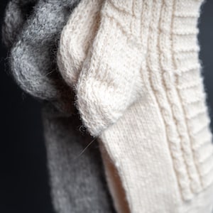 Pode incluir: Close-up de três meias de malha em tons de cinza e creme. A meia creme apresenta um design de malha de cabo. As meias são feitas de um fio macio e texturizado, perfeito para o frio.