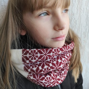Könnte beinhalten: Ein gestrickter Cowl-Schal mit Fair-Isle-Muster. Der Schal hat ein weißes und bordeauxfarbenes geometrisches Design. Der Cowl wird um den Hals getragen und mit einer schwarzen Jacke kombiniert. Ein gemütliches Winteraccessoire.