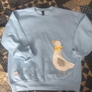 May include: Light blue sweatshirt with a white goose applique. The goose has a yellow bow and a gray patterned wing. The sweatshirt has a crew neck and long sleeves. A small tag is visible on the lower left side.