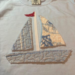 May include: A light blue t-shirt featuring a sailboat appliqué made of quilted fabric patches. The boat hull and sails are crafted from light blue and white patterned fabric, with a small red patterned flag at the top of the white mast.