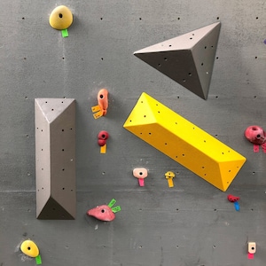 May include: A close-up of a gray wall with various colored climbing holds. The holds are made of different materials, including plastic and wood. The holds are arranged in a random pattern on the wall.
