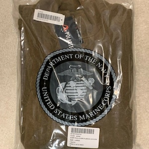 May include: A brown fleece pullover shirt with a zipper. The shirt has the United States Marine Corps logo on the front. The shirt is packaged in a plastic bag with a label that says "CAGE: 4Z046 SHIRT PULLOVER FLEECE COYOTE SIZE: LARGE 1 EACH SPM1C1-07-0-N023 A"