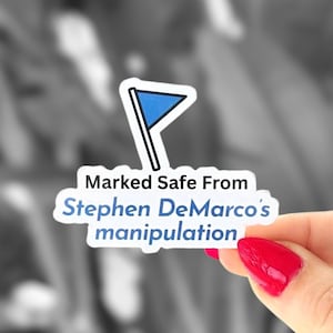 May include: A white sticker with a blue flag design and the text "Marked Safe From Stephen DeMarco's manipulation". The sticker is held by a hand with red nail polish.