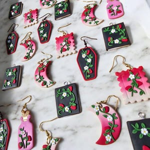 以下が含まれることがあります： 棺、三日月、本、ボトルなど、さまざまな形とデザインを特徴とする、手作りポリマークレイのイヤリングの詰め合わせ。各作品は、イチゴと白い花のつるで飾られています。