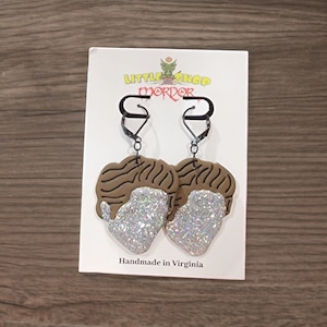 May include: A pair of silver glitter earrings shaped like a man's face with brown hair. The earrings are on a white card with the text "Little Shop of Horrors" and "Handmade in Virginia".