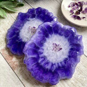 May include: Two resin coasters in shades of purple and white, featuring a floral pattern. The coasters have a scalloped edge and a white central area with small amethyst crystals. A small white dish with amethyst crystals is also present.