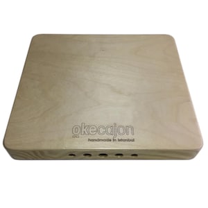 May include: A light-colored wooden cajon drum with rounded corners. The text "okecajon" and "handmade in Istanbul" are printed on the front. The drum has several sound holes near the bottom edge.