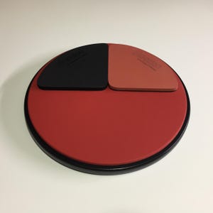 May include: A red, black, and brown practice drum pad with a black rim. The pad has three different surfaces: a black "Soft" surface, a brown "Medium" surface, and a red "Hard" surface.
