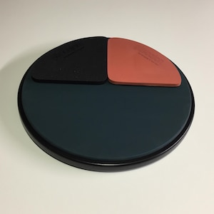 May include: A black, red, and green practice drum pad with a black rim. The pad has three different surfaces for practicing different drum sounds.