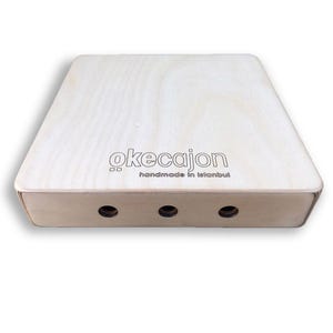 May include: A light wood cajon with the text "okecajon" and "handmade in Istanbul" on the front. The cajon has three sound holes.