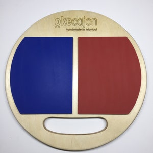 May include: A round wooden cajon with a blue and red playing surface. The cajon has a handle and the text "okecajon handmade in Istanbul" is printed on the front.