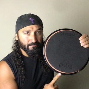 May include: A person holding a black and brown round drum pad with the text "okecajon" printed on it. The drum pad has a small graphic of a person playing drums.