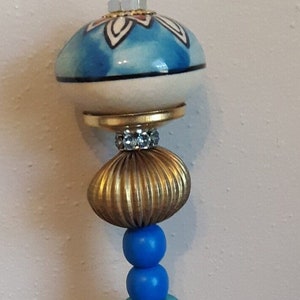 May include: A decorative beaded ornament with a blue and white ceramic top. The ornament features various beads in blue, gold, brown, clear, and purple. The ornament is hanging against a neutral background.