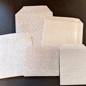 May include: Five white paper gift bags with a black and white geometric pattern. The bags are arranged in a stack with the largest bag on the bottom and the smallest bag on top.