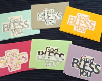 May include: A collection of colorful greeting cards with the message "God Bless You" in a cross-shaped design. The cards are in various colors, including pink, green, yellow, and blue, with patterned text.
