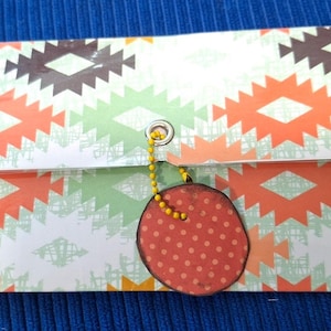 May include: A white envelope with a colorful geometric pattern. The envelope has a red circle with white polka dots attached to it with a yellow chain.