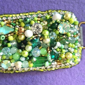 May include: A rectangular, beaded accessory with a gold-colored ring. The surface is densely covered with green, white, and clear beads, crystals, and rhinestones. The edges are outlined with small gold and silver beads.