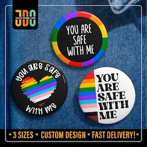 Puede incluir: Tres chapas redondas con el texto "YOU ARE SAFE WITH ME". Una chapa tiene fondo negro con borde arcoíris, otra fondo negro con un corazón arcoíris y la última fondo blanco con una franja vertical de colores del arcoíris.