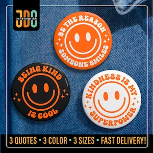 May include: Three round button pins with different messages and smiley faces. The first pin is orange with white text and a white smiley face. The text reads "Be the reason someone smiles". The second pin is black with orange text and a orange smiley face. The text reads "Being kind is cool". The third pin is white with orange text and an orange smiley face. The text reads "Kindness is my superpower".