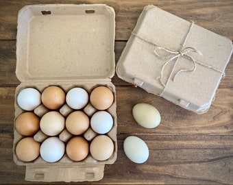 May include: An open cardboard egg carton filled with a dozen eggs in varying shades of brown and white. A closed carton tied with twine sits beside it, along with two loose eggs. The eggs rest on a wooden surface.